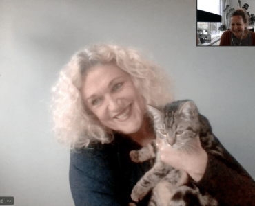 Mirjam Kaijer glimlacht, zij draagt een zwart trui en houdt een getijgerde kat vast. Haar blonde krullen lichten op door daglicht van een raam aan de zijkant. Inzet podcasthost Jolanda Gooiker